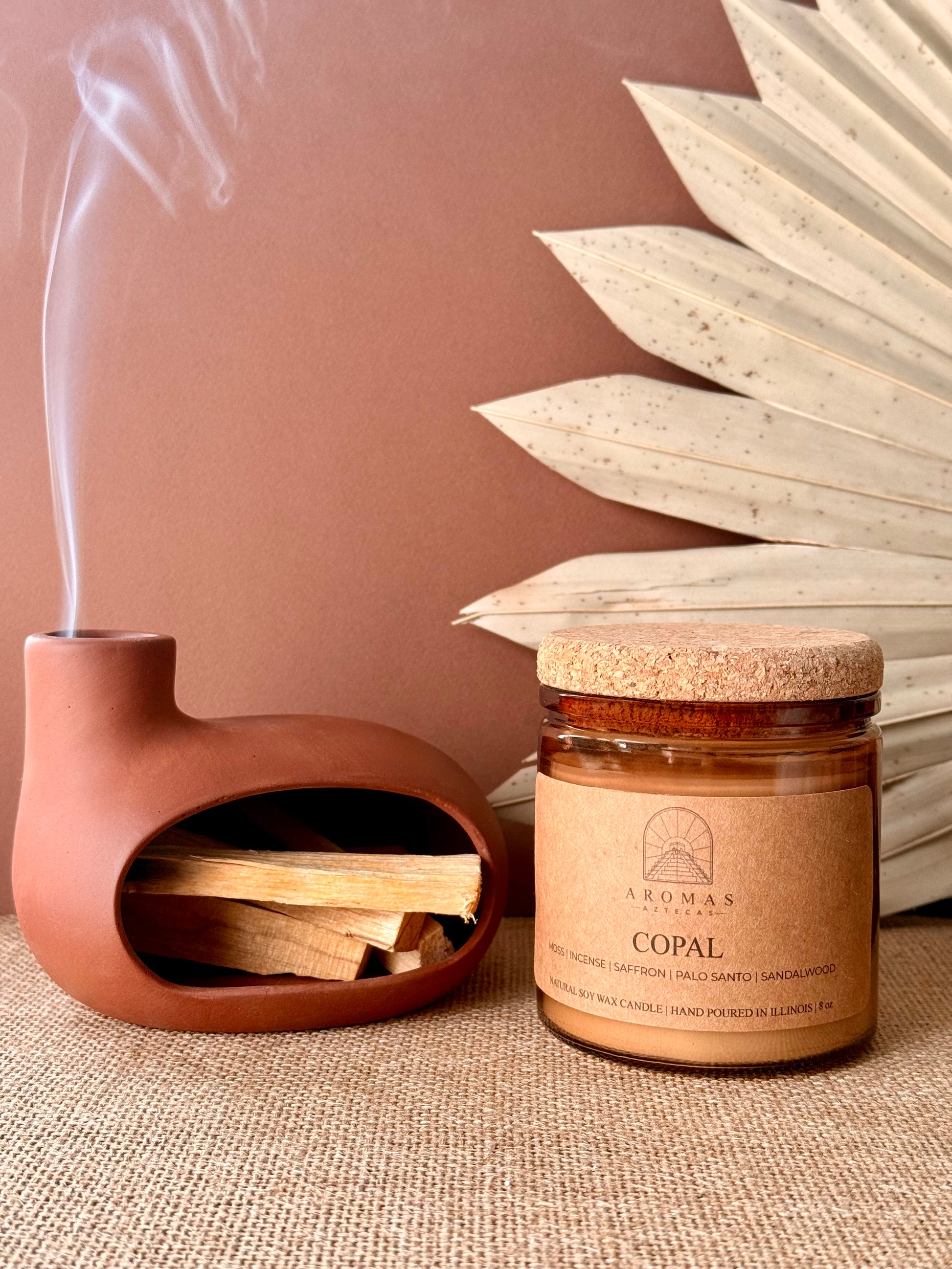 Aromas Aztecas Copal soy candle in an amber glass vessel with a cork lid on a woven textured surface, dried palm leaf décor, terracotta pottery, and smoldering palo santo.