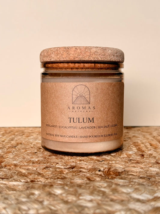 Aromas Aztecas Tulum soy candle in an amber glass vessel with a cork lid on a woven textured surface.