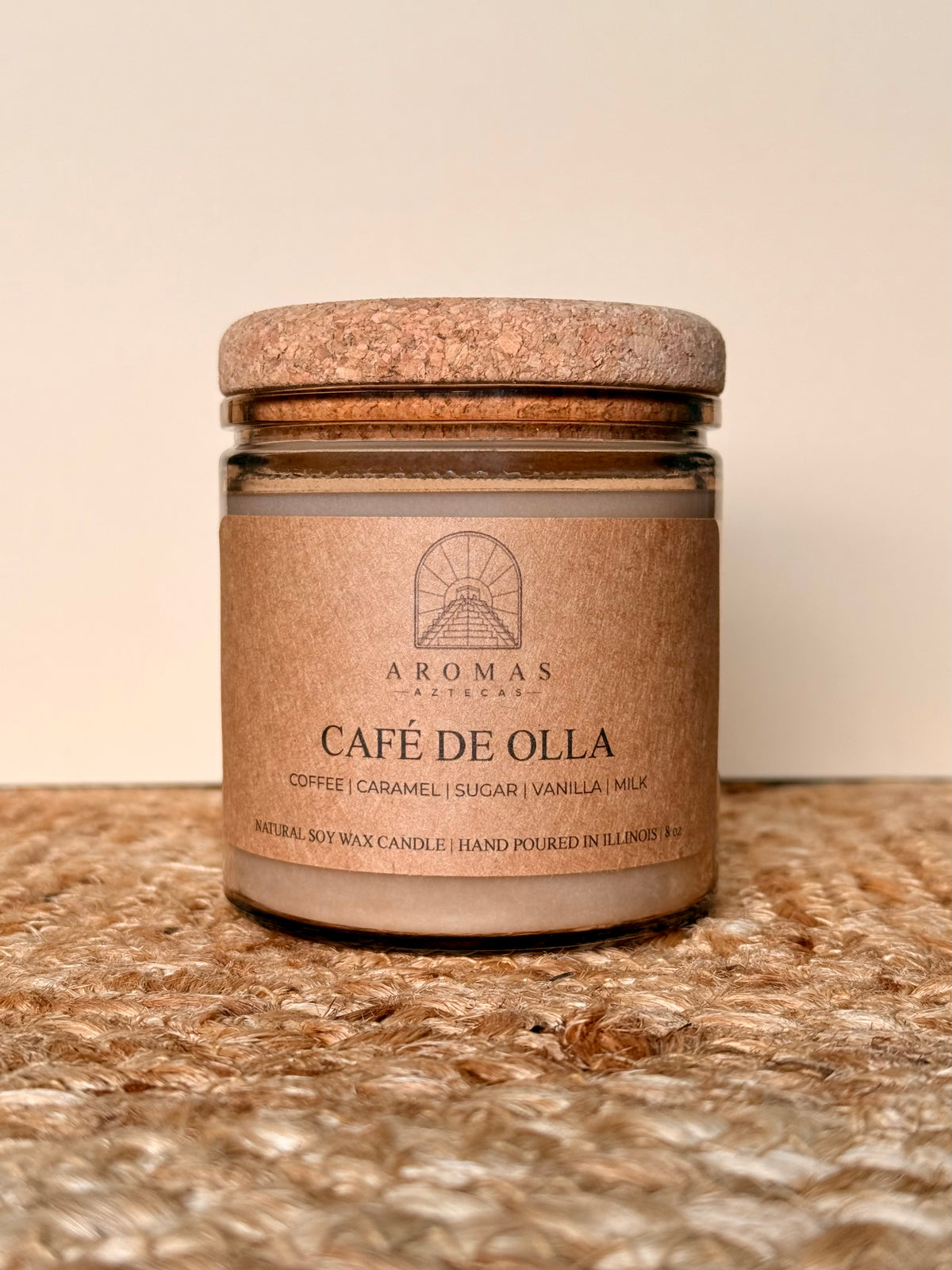 Aromas Aztecas Café de Olla soy candle in an amber glass vessel with a cork lid on a woven textured surface.