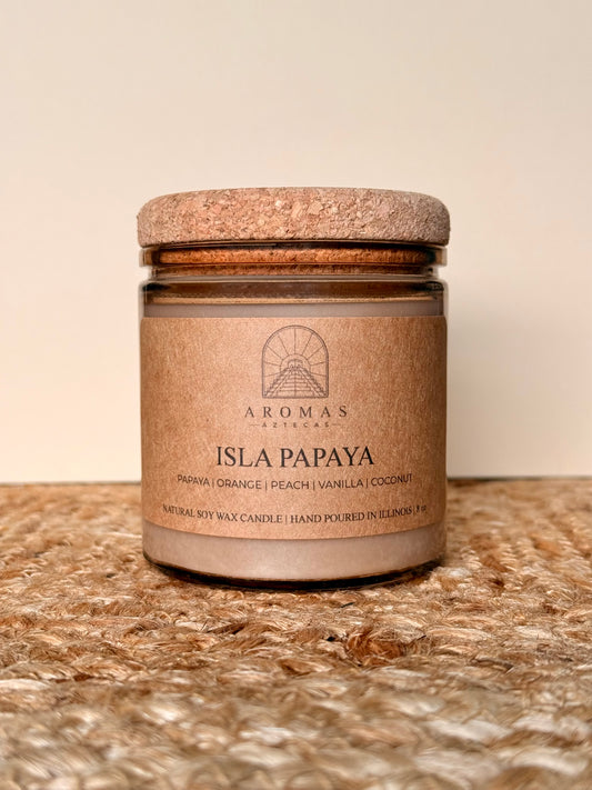 Aromas Aztecas Isla Papaya soy candle in an amber glass vessel with a cork lid on a woven textured surface.