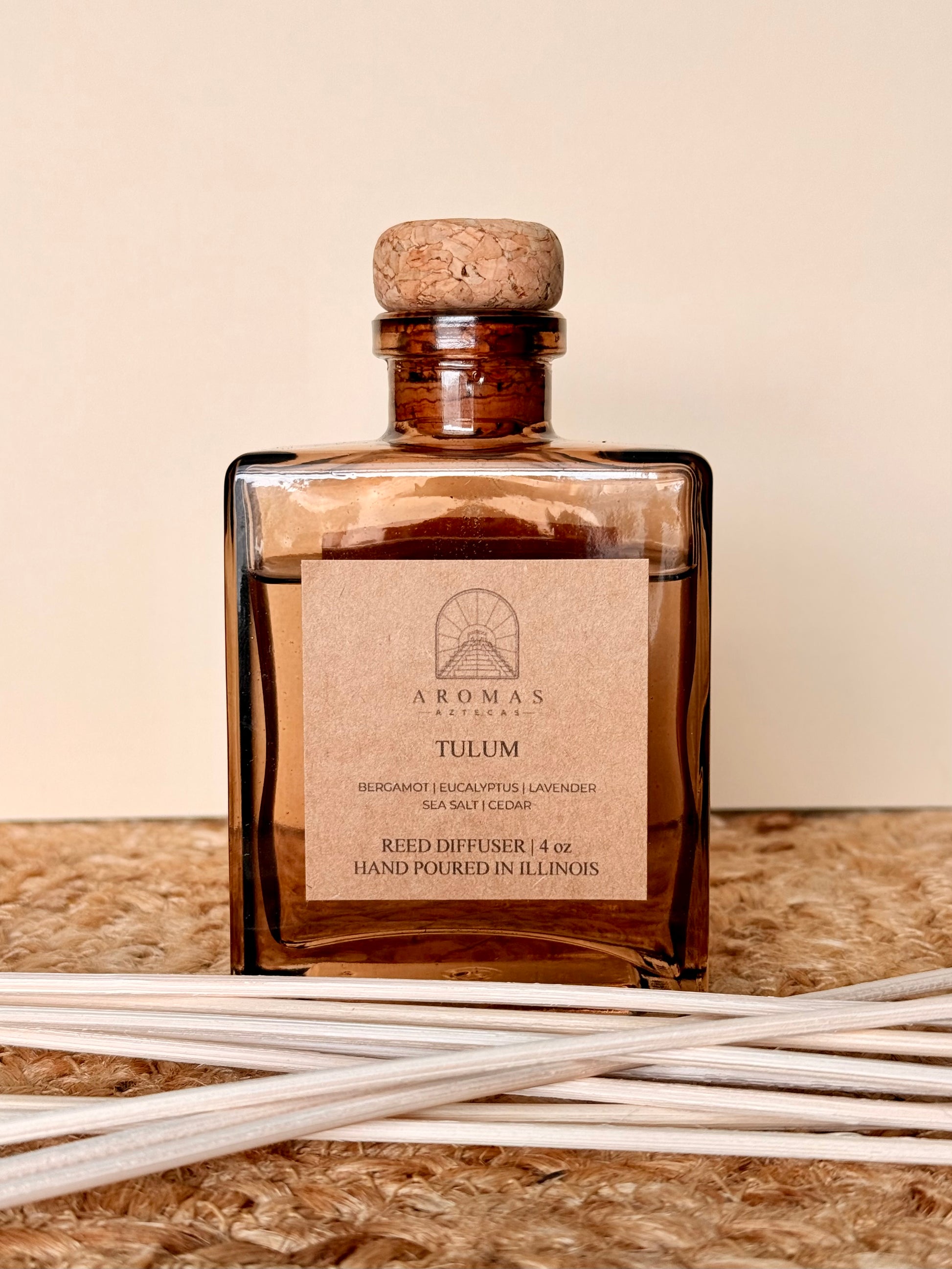 Aromas Aztecas Tulum reed diffuser in an amber glass bottle with natural reeds on a woven textured surface.