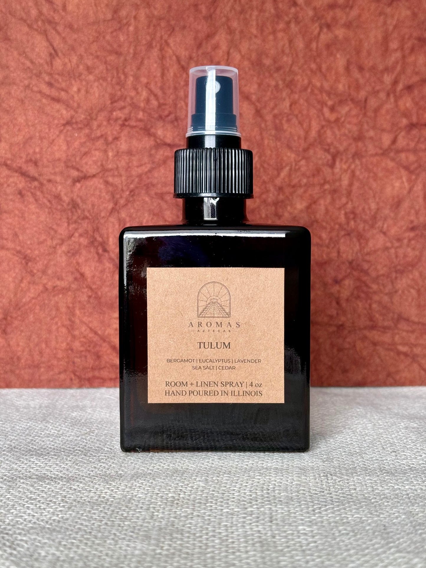 Aromas Aztecas Tulum room and linen spray in an amber glass bottle on a woven textured surface with a burgundy textured backdrop.