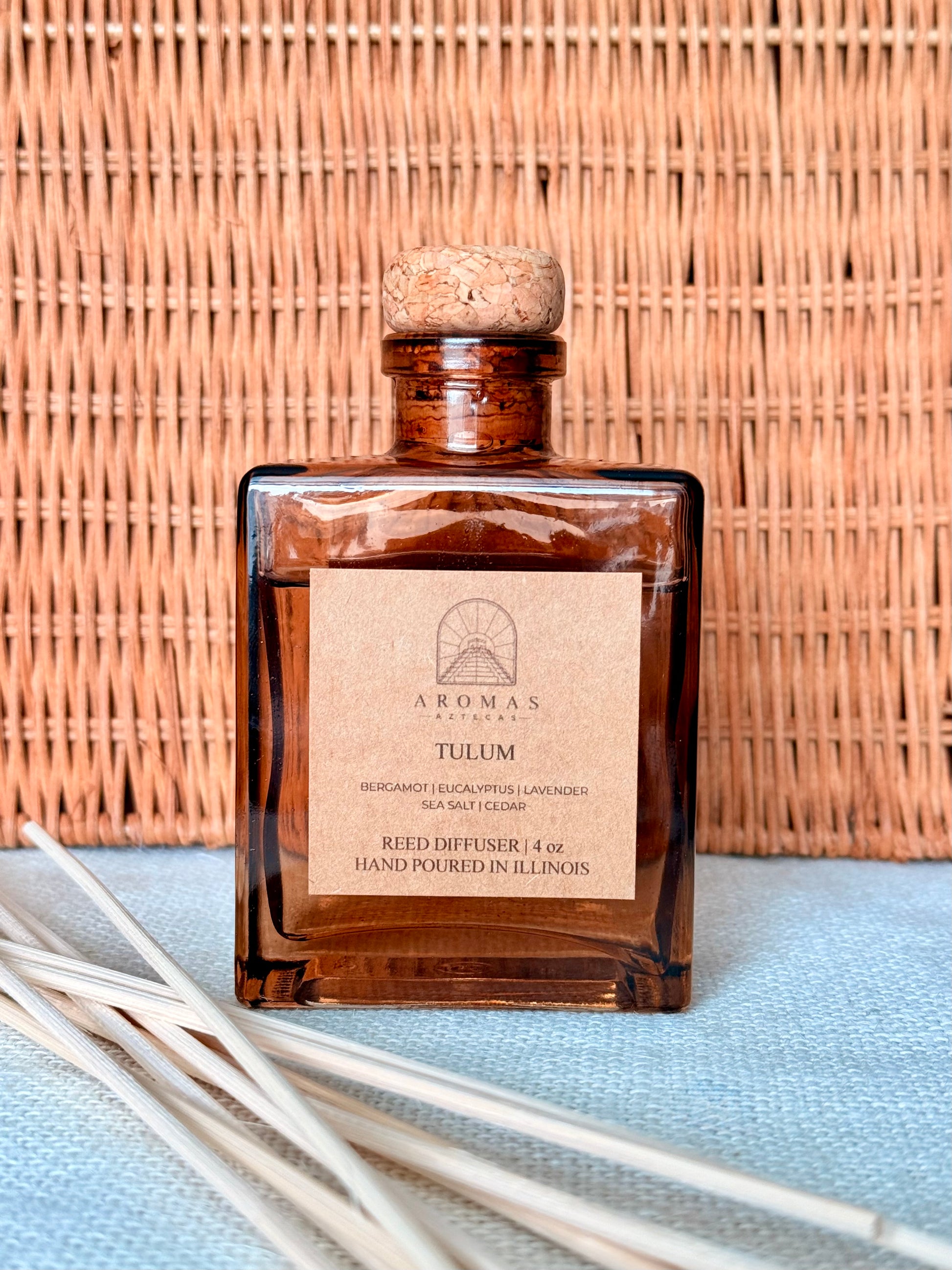 Aromas Aztecas Tulum reed diffuser in an amber glass bottle with natural reeds against a woven textured backdrop.