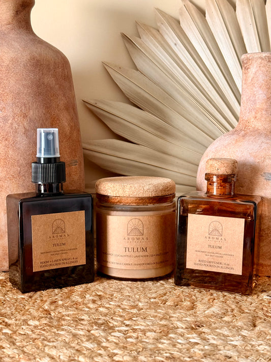 Aromas Aztecas Tulum room and linen spray, soy candle, and reed diffuser in amber glass vessels on a woven textured surface with terracotta pottery and dried palm leaf décor.