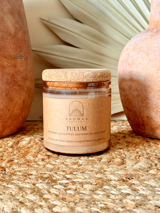 Aromas Aztecas Tulum soy candle in an amber glass vessel with a cork lid on a woven textured surface with terracotta pottery and dried palm leaf décor.