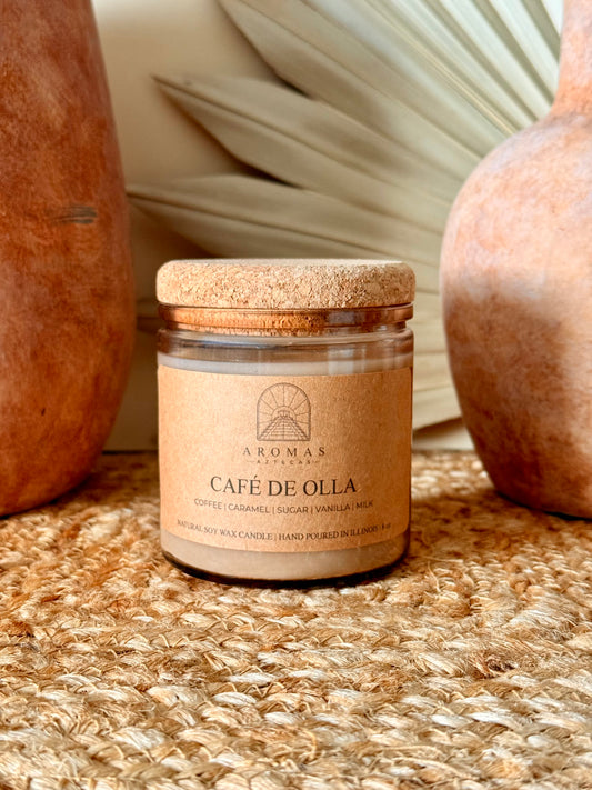 Aromas Aztecas Café de Olla soy candle in an amber glass vessel with a cork lid on a woven textured surface with terracotta pottery and dried palm leaf décor.