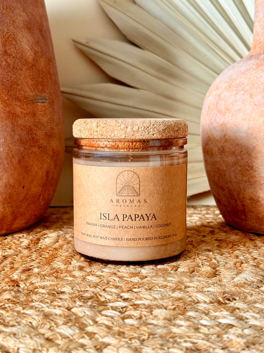 Aromas Aztecas Isla Papaya soy candle in an amber glass vessel with a cork lid on a woven textured surface with terracotta pottery and dried palm leaf décor.