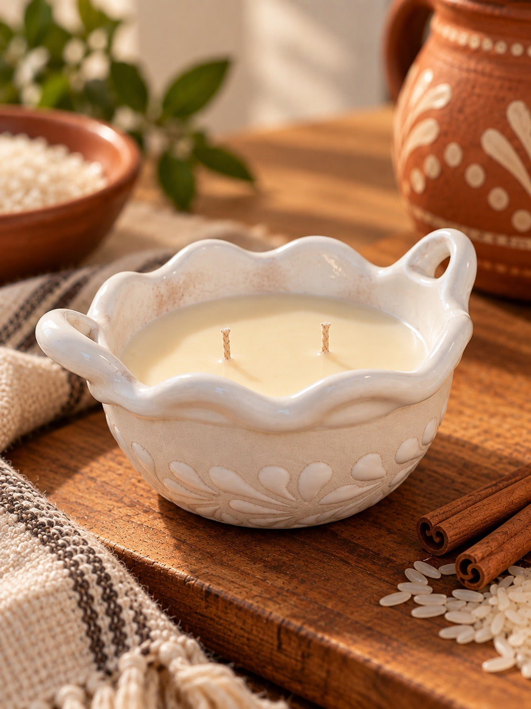Arroz con Leche handcrafted white talavera cazuelita candle with two wicks on a rustic wooden table, surrounded by rice, cinnamon sticks, terracotta pottery, and a woven textile.