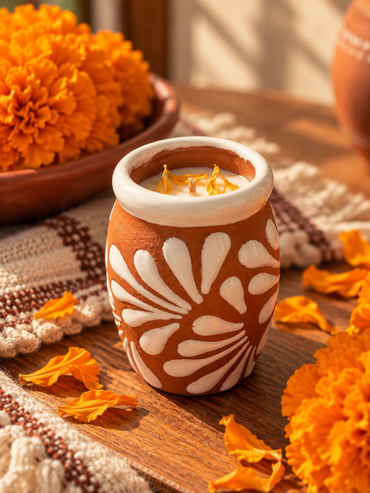 A handcrafted terracotta candle vessel filled with white wax layered with vibrant orange marigold petals and surrounded by marigold petals and flowers on a rustic wooden table and a textured textile.