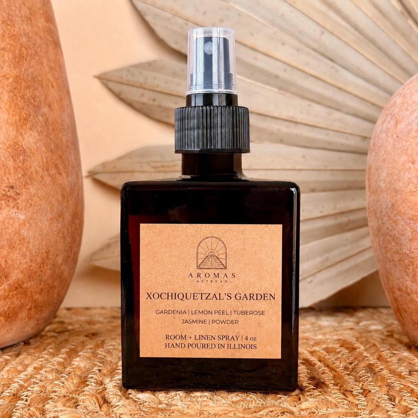 Aromas Aztecas Xochiquetzal's Garden room and linen spray in an amber glass bottle on a woven textured surface with terracotta pottery and dried palm leaf décor.