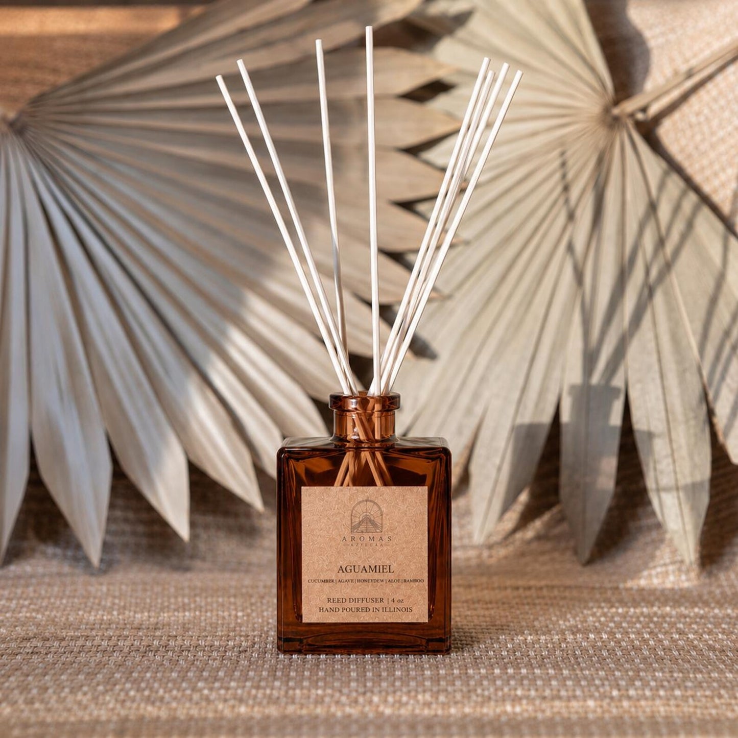 Aromas Aztecas Aguamiel reed diffuser in an amber glass bottle with natural reeds on a woven textured surface and dried palm leaf décor.