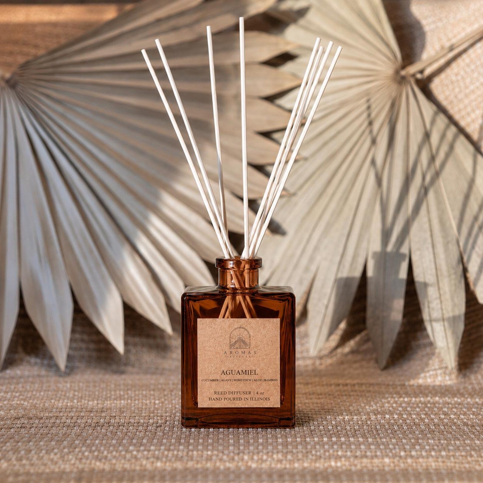 Aromas Aztecas Aguamiel reed diffuser in an amber glass bottle with natural reeds on a woven textured surface and dried palm leaf décor.