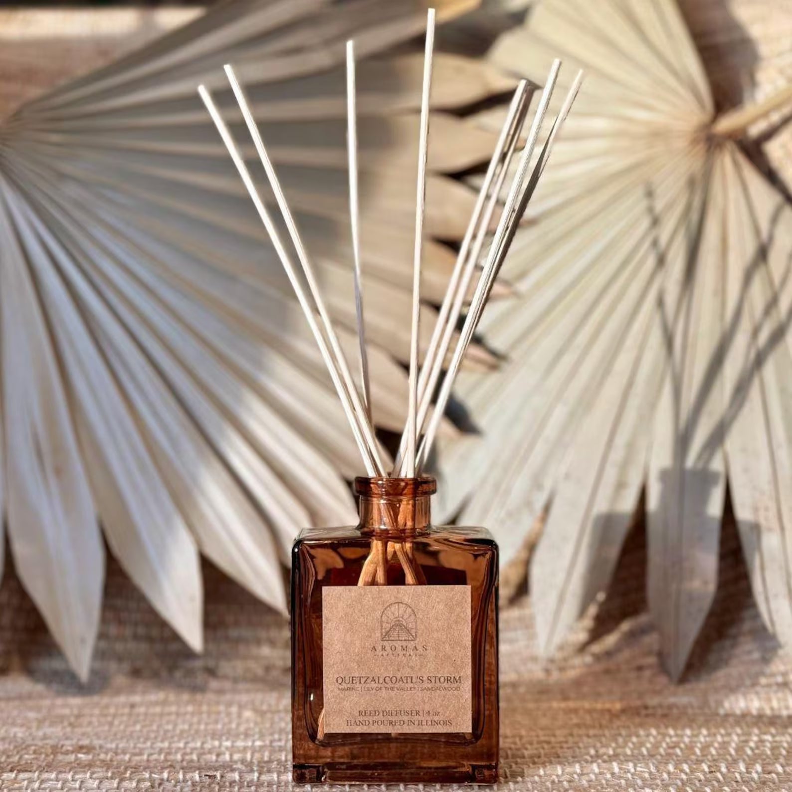 Aromas Aztecas Quetzalcoatl's Storm reed diffuser in an amber glass bottle with natural reeds on a woven textured surface and dried palm leaf décor.