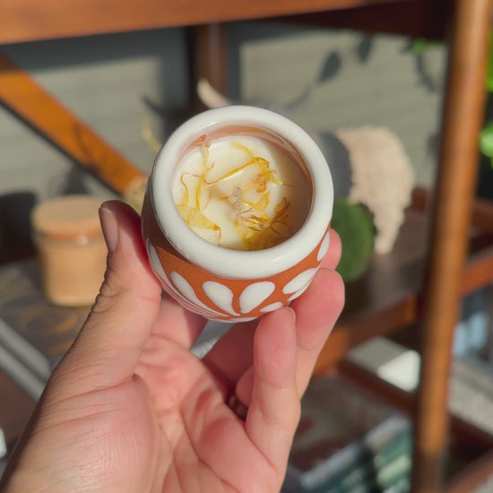 Video of a hand holding a small terracotta colored ceramic Cempasúchil velita with a white floral pattern and white rim, photographed in warm natural light with a blurred bookshelf in the background. The inside of the velita is visible showing the wax, wick, and marigold petals.