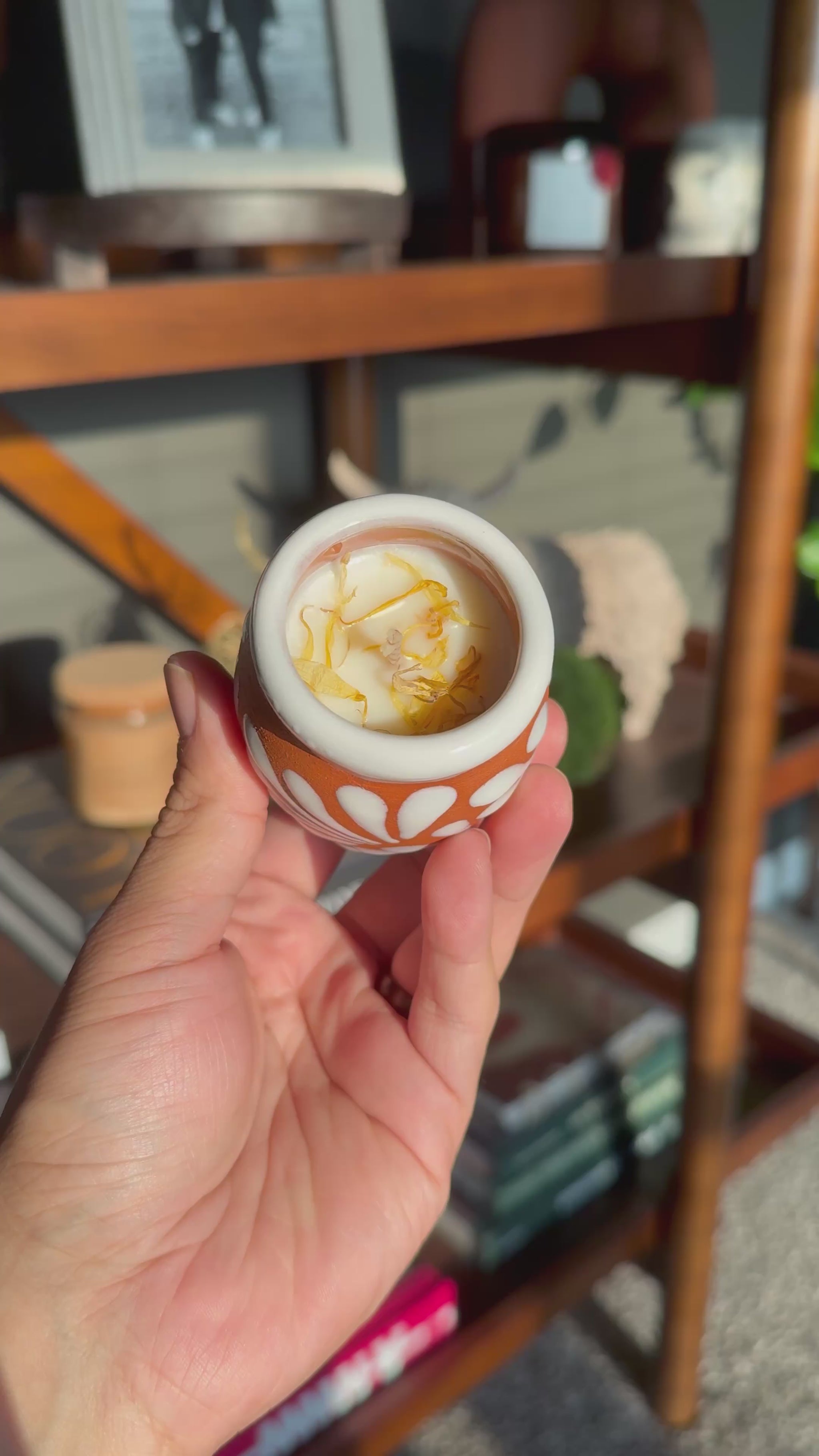 Video of a hand holding a small terracotta colored ceramic Cempasúchil velita with a white floral pattern and white rim, photographed in warm natural light with a blurred bookshelf in the background. The inside of the velita is visible showing the wax, wick, and marigold petals.